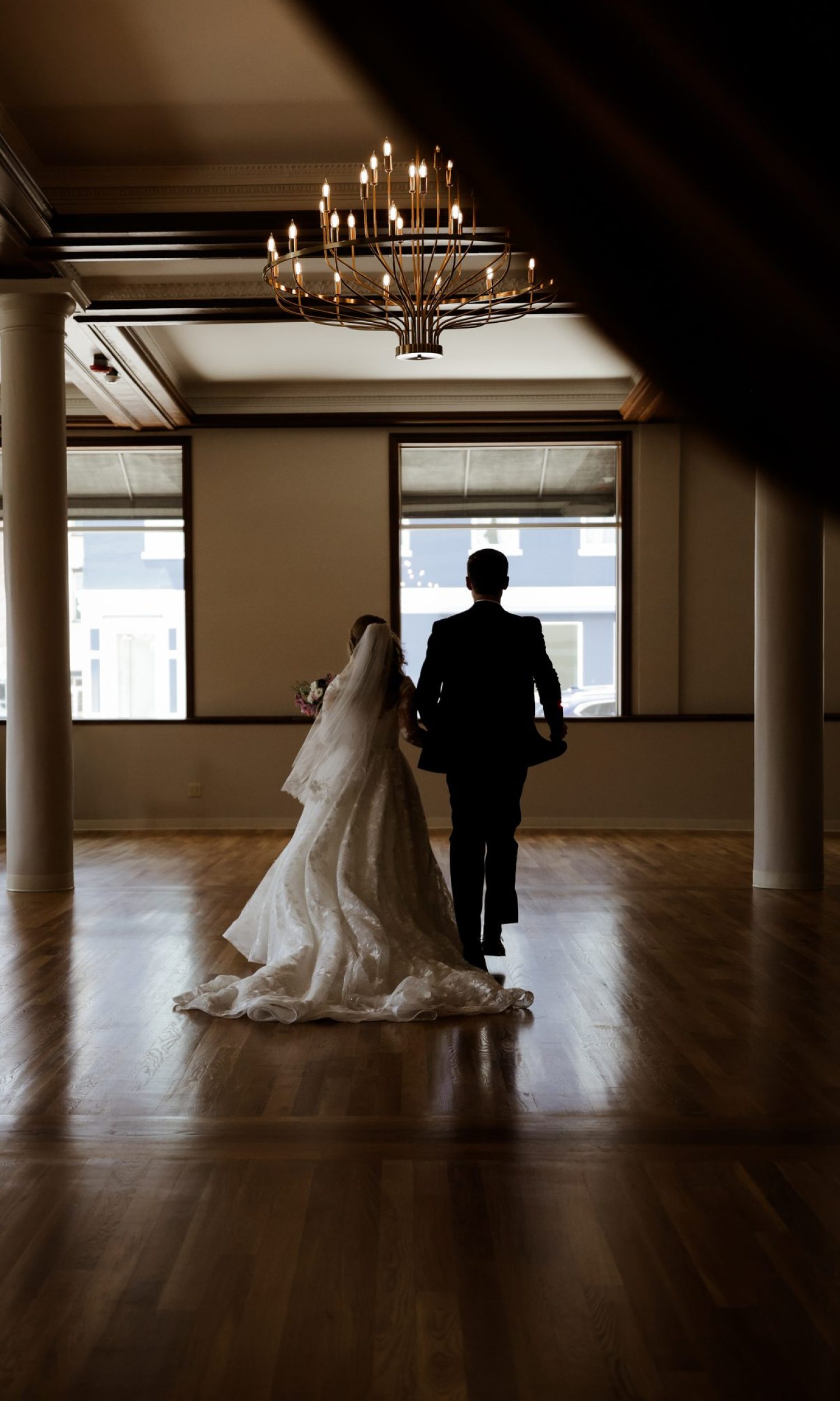 Couple walking across the hardwood floor of The Event Vault main hall under the chandelier